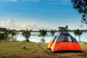 Orange camping tent set up beside a calm lake under blue sky